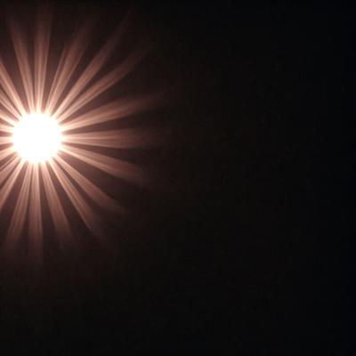 The primary subject of the image is a bright light source, possibly a sun or a light bulb, shining through a dark background. The light source appears to be the main focus of the image, creating a contrast between the bright light and the surrounding darkness. The visual style of the image is a close-up of the light source, emphasizing its intensity and creating a dramatic effect. The colors and mood of the image are predominantly dark, with the bright light source serving as the only source of illumination, giving